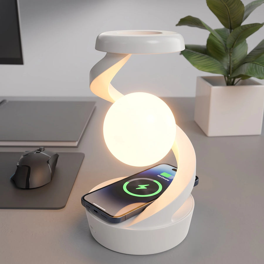 A white modern spiral-shaped desk lamp with a built-in charging base, shown glowing on a gray desk next to a smartphone. A potted plant and a laptop are visible in the background.